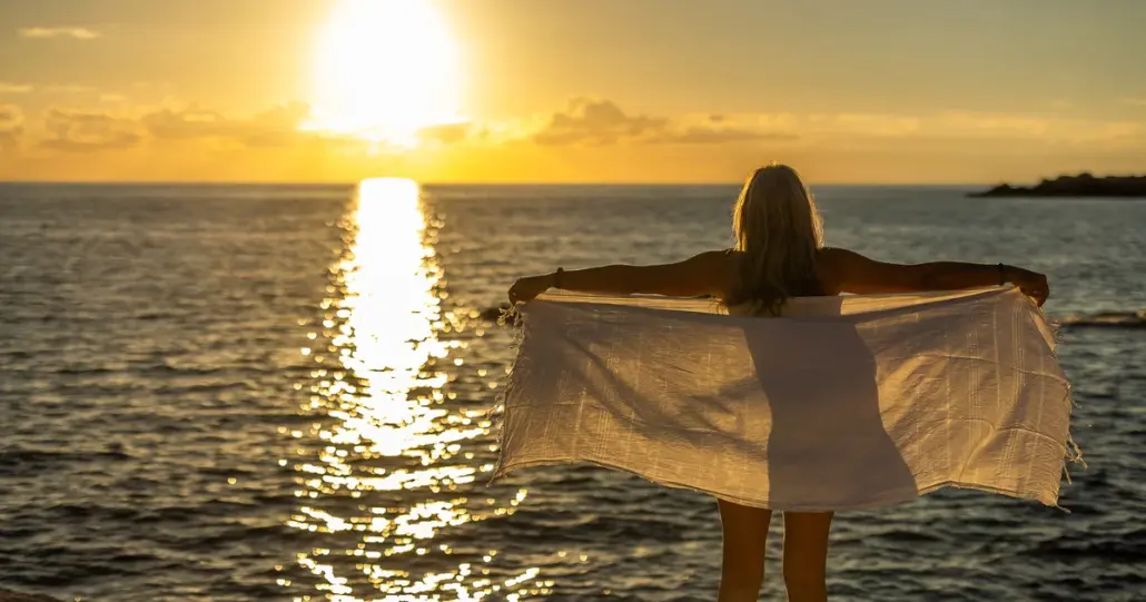 Eine Frau steht am Meer mit dem Rücken zur Kamera und hält im Gegenlicht der tief stehenden Sonne ein helles Tuch mit ausgebreiteten Armen.