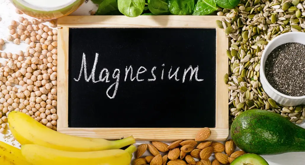 Kleine Schiefertafel mit der Aufschrift „Magnesium”, umgeben von Spinat, Kichererbsen, Kürbis- und Sonnenblumenkernen, Chiasamen, Mandeln, Bananen und Avocado.